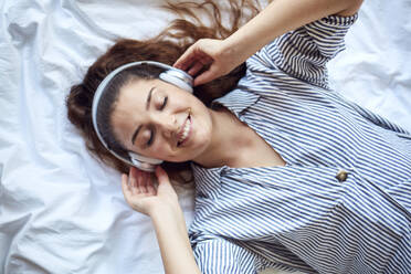 Portrait of happy young woman with eyes closed lying on bed listening music with headphones - JHAF00110