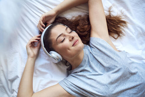 Porträt einer glücklichen jungen Frau mit geschlossenen Augen, die auf dem Bett liegt und mit Kopfhörern Musik hört - JHAF00112