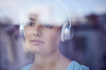 Porträt einer jungen Frau, die mit Kopfhörern Musik hört und aus dem Fenster schaut - JHAF00116