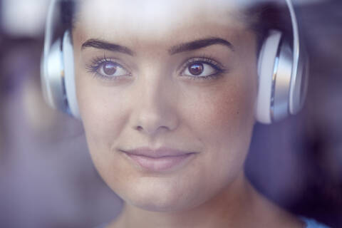 Porträt einer jungen Frau, die mit Kopfhörern Musik hört und aus dem Fenster schaut, lizenzfreies Stockfoto