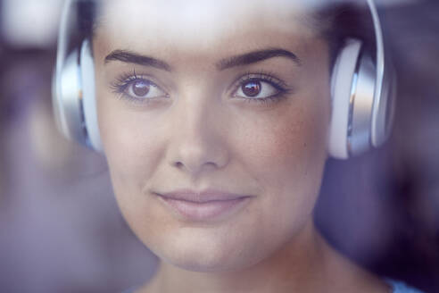 Porträt einer jungen Frau, die mit Kopfhörern Musik hört und aus dem Fenster schaut - JHAF00117