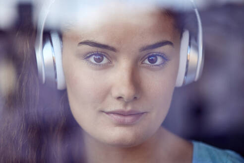 Porträt einer jungen Frau hinter einer Fensterscheibe, die mit Kopfhörern Musik hört - JHAF00118