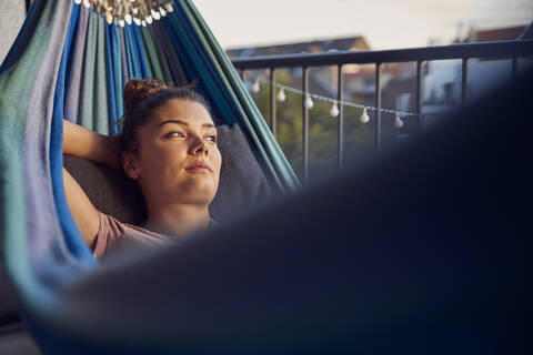 Porträt einer nachdenklichen jungen Frau, die in einer Hängematte auf einem Balkon liegt und in die Ferne schaut, lizenzfreies Stockfoto