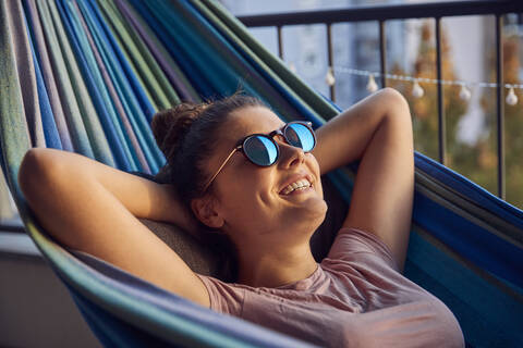 Porträt einer glücklichen jungen Frau mit Sonnenbrille in der Hängematte auf dem Balkon, lizenzfreies Stockfoto