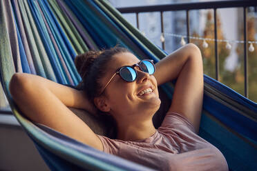Portrait of happy young woman with sunglasses lying on hammock on balcony - JHAF00126