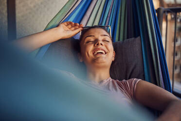 Portrait of happy young woman lying on hammock on balcony in the evening - JHAF00127