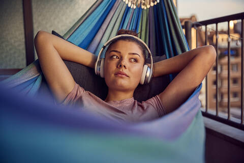 Porträt einer jungen Frau, die in einer Hängematte auf dem Balkon liegt und mit Kopfhörern Musik hört, lizenzfreies Stockfoto