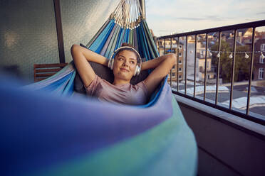 Portrait of smiling young woman lying on hammock on balcony listening music with headphones - JHAF00131