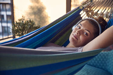 Portrait of smiling young woman lying on hammock on balcony listening music with headphones - JHAF00133