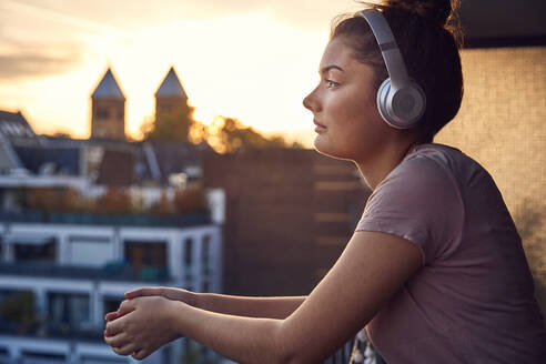 Junge Frau hört Musik mit Kopfhörern auf dem Balkon bei Sonnenuntergang - JHAF00134