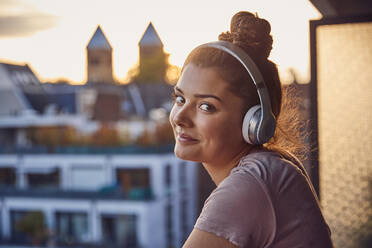 Portrait of smiling young woman with bun listening music with headphones on balcony in the evening - JHAF00135