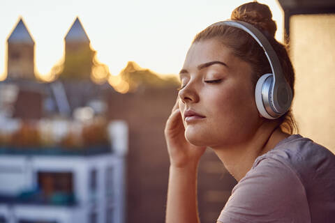 Porträt einer jungen Frau mit geschlossenen Augen, die mit Kopfhörern auf dem Balkon bei Sonnenuntergang Musik hört, lizenzfreies Stockfoto