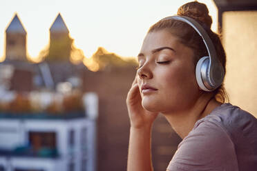 Porträt einer jungen Frau mit geschlossenen Augen, die mit Kopfhörern auf dem Balkon bei Sonnenuntergang Musik hört - JHAF00136