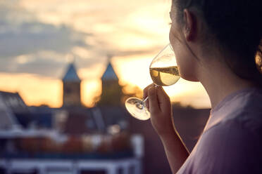 Junge Frau auf dem Balkon trinkt ein Glas Weißwein bei Sonnenuntergang - JHAF00138