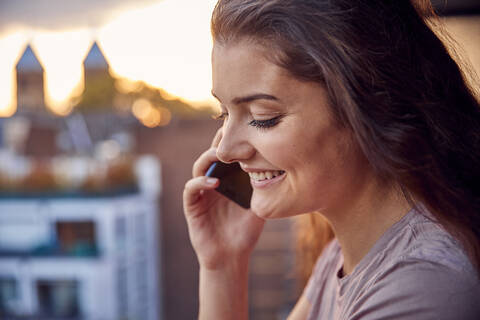 Lächelnde junge Frau beim Telefonieren auf dem Balkon bei Sonnenuntergang, lizenzfreies Stockfoto