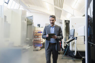 Mature businessman walking on production floor of factory, using digital tablet - DIGF09843