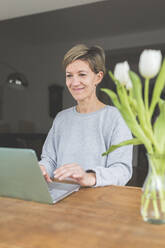 Mature woman working from home, using laptop on table with flowers - ASCF01228