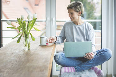 Reife Frau, die von zu Hause aus arbeitet, Laptop benutzt und im Schneidersitz auf einer Bank sitzt - ASCF01246