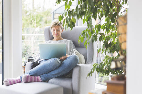 Ältere Frau, die von zu Hause aus arbeitet, mit Laptop, im Sessel sitzend - ASCF01249