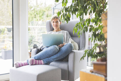 Ältere Frau, die von zu Hause aus arbeitet, mit Laptop, im Sessel sitzend - ASCF01250