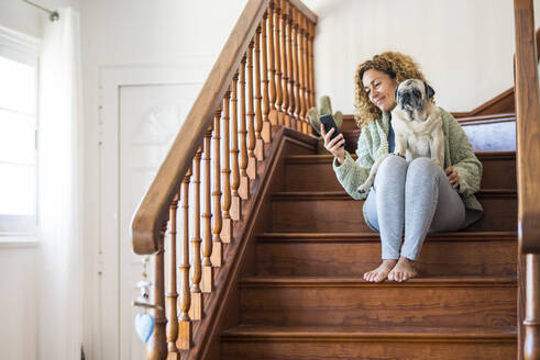 Woman sitting on stairs, using smartphone with pug on her lap - SIPF02154