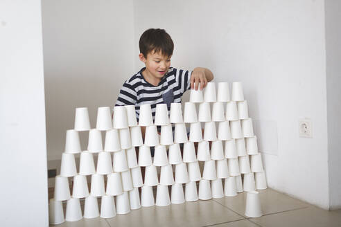 Lächelnder Junge baut zu Hause eine Wand aus weißen Pappbechern - HMEF00890