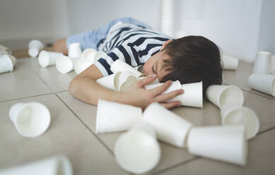 Little boy lying on the floor between white paper cups - HMEF00892