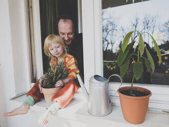 Father with daughter sitting on windowsill by potted plant - IHF00332