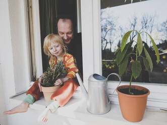 Vater mit Tochter auf der Fensterbank neben einer Topfpflanze sitzend - IHF00332