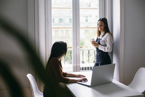 Geschäftsfrau trinkt Kaffee, während ein Kollege im Büro am Laptop arbeitet, lizenzfreies Stockfoto