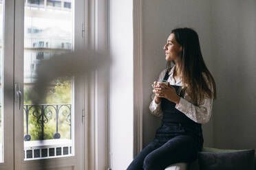 Nachdenkliche Geschäftsfrau schaut durch das Fenster, während sie im Büro einen Kaffee trinkt - ABZF03087