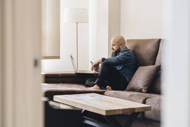 Geschäftsmann mit Laptop, der ein Mobiltelefon benutzt, während er zu Hause auf dem Sofa sitzt - DGOF00892