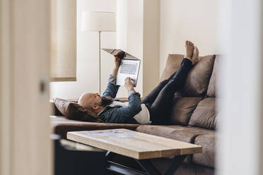 Bored man using laptop while lying on sofa in living room at home - DGOF00893