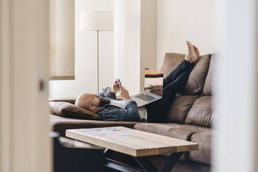 Geschäftsmann mit Laptop hört Musik über sein Smartphone, während er zu Hause auf dem Sofa liegt - DGOF00895