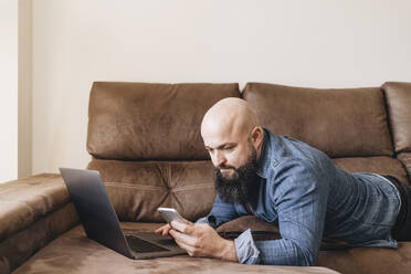 Geschäftsmann mit Laptop und Mobiltelefon, während er zu Hause auf dem Sofa liegt - DGOF00898