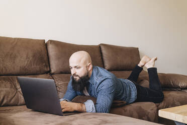 Geschäftsmann, der einen Laptop benutzt, während er zu Hause auf dem Sofa im Wohnzimmer liegt - DGOF00899