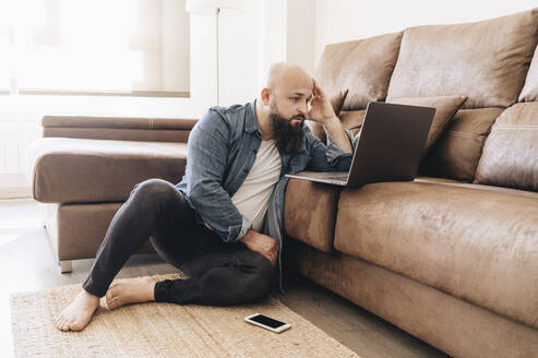 Seriöser Geschäftsmann, der einen Laptop benutzt, während er zu Hause auf dem Boden neben dem Sofa sitzt - DGOF00900