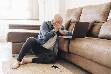 Müder Geschäftsmann mit Laptop, der das Gesicht bedeckt, während er zu Hause im Wohnzimmer sitzt - DGOF00901