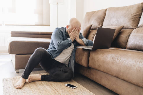 Tired businessman with laptop covering face while sitting in living room at home - DGOF00901