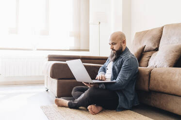 Geschäftsmann mit rasiertem Kopf arbeitet am Laptop, während er zu Hause auf dem Boden vor dem Sofa sitzt - DGOF00903