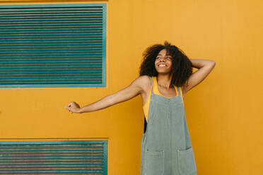 Portrait of happy young woman wearing overalls in front of yellow wall - TCEF00474