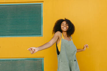 Portrait of happy young woman wearing overalls in front of yellow wall - TCEF00475