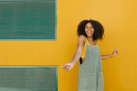 Porträt einer fröhlichen jungen Frau in Latzhose vor einer gelben Wand, lizenzfreies Stockfoto