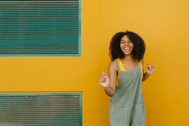 Portrait of happy young woman wearing overalls in front of yellow wall - TCEF00477