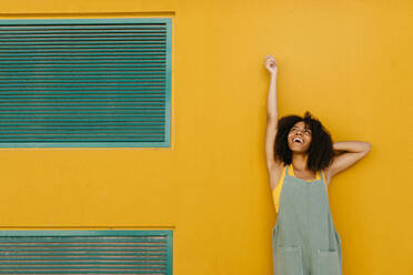 Portrait of happy young woman wearing overalls in front of yellow wall - TCEF00478