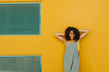 Portrait of happy young woman wearing overalls in front of yellow wall - TCEF00479