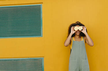 Happy young woman wearing overalls in front of yellow covering her eyes with lemons - TCEF00480