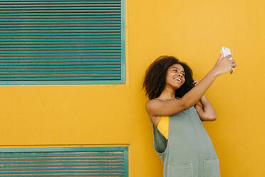Porträt einer glücklichen jungen Frau in Latzhose vor einer gelben Wand, die ein Selfie macht - TCEF00484