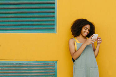 Lächelnde junge Frau in Latzhose vor einer gelben Wand, die ein Smartphone benutzt - TCEF00487