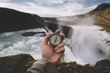 Island, Hand einer Person, die einen Navigationskompass gegen den plätschernden Wasserfall Gullfoss hält - DAMF00366